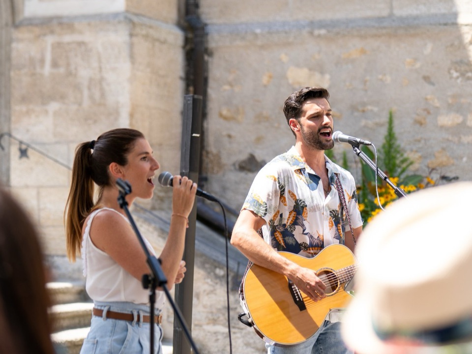 Yann à la guitare et une chanteuse au micro, en plein air devant une église, le public au premier plan