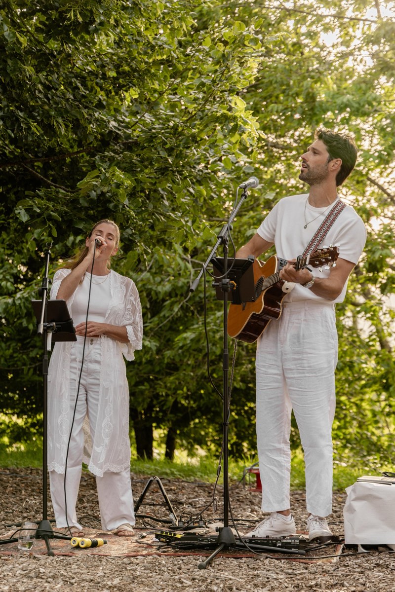 Yann Wellhauser à la guitare et une chanteuse, tout en blanc, en concert sous les arbres en fin de journée