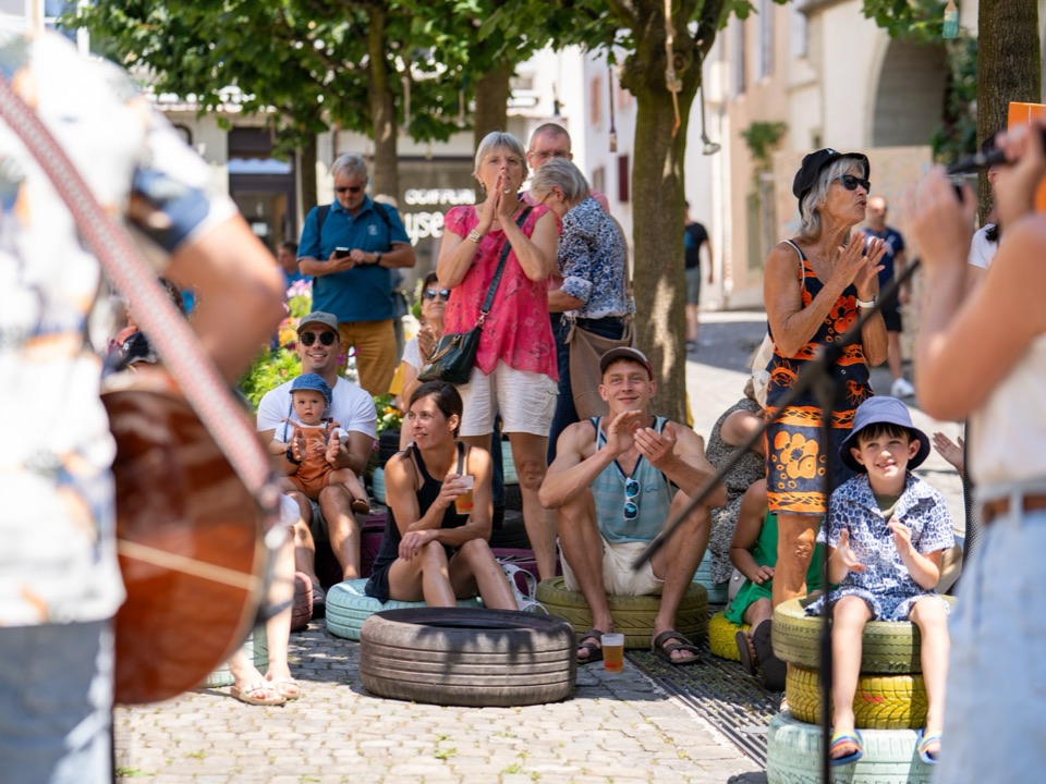 Des familles assises dans une rue piétonne qui applaudissent un concert, des enfants parmi elles