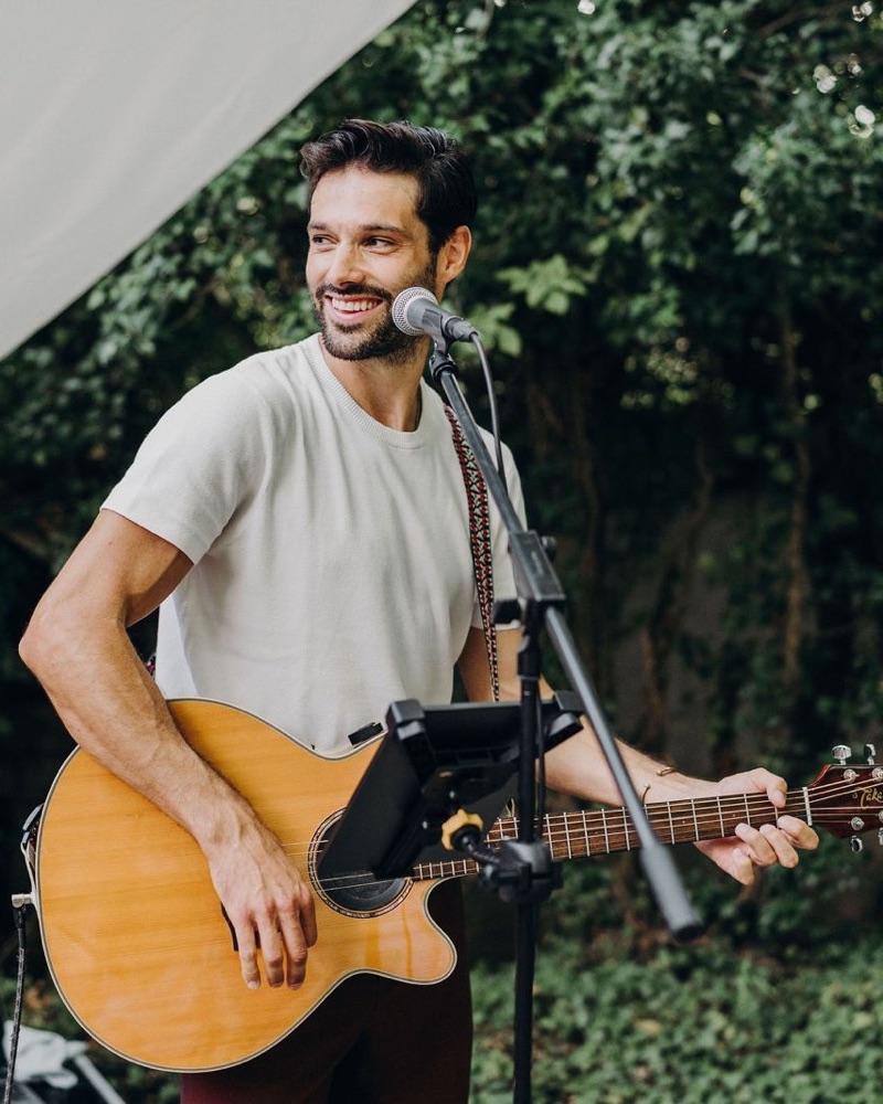 Yann Wellhauser qui sourit en jouant de la guitare au micro, en t-shirt blanc, dans un jardin sous une tente